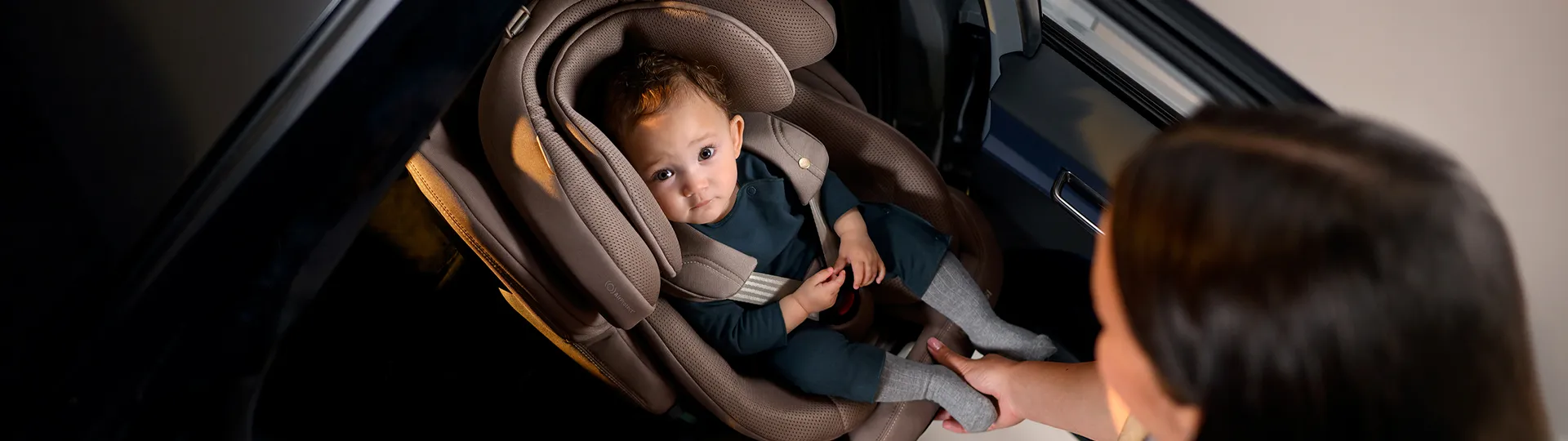 Ein Baby in einem braunen Kindersitz blickt nach oben, während ein Erwachsener dem Kind in einem Fahrzeug einen kleinen Gegenstand überreicht.