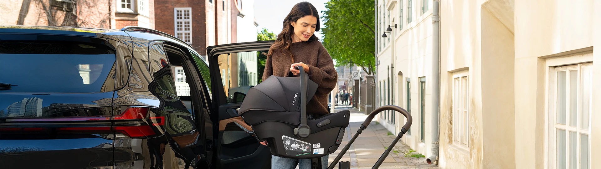 Eine Frau stellt einen schwarzen Babysitz in einen Kinderwagen neben einem geparkten Auto auf einer sonnigen Straße in der Stadt.