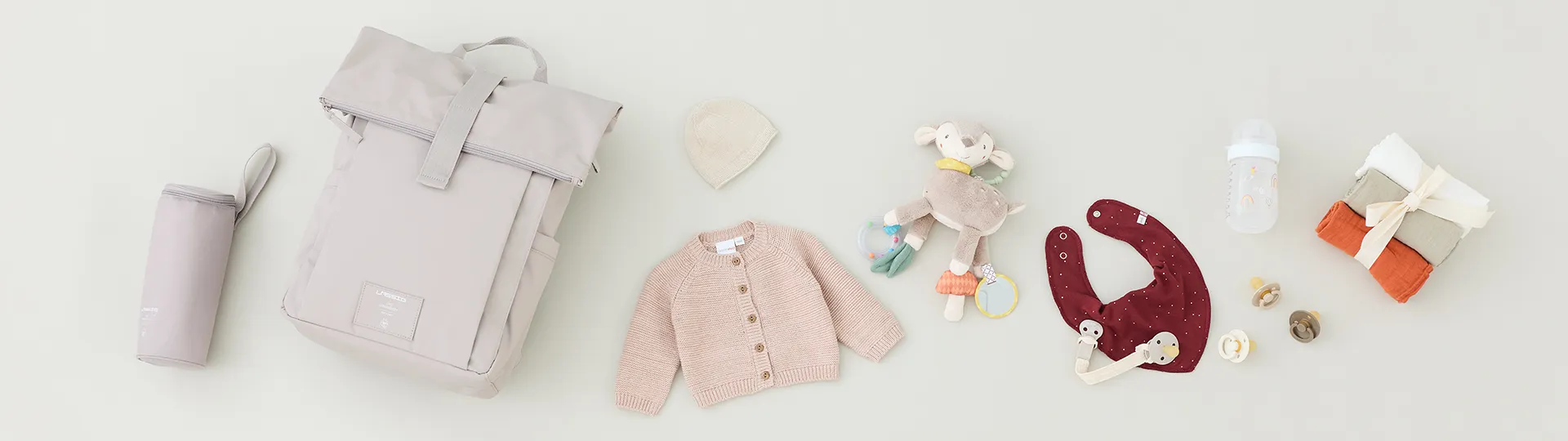 Zur Babyausstattung gehören ein beiger Rucksack, eine Strickjacke, eine Mütze, ein Plüschtier, Lätzchen, eine Flasche, ein Stapel Stofftücher und Schnuller, die auf einem hellen Hintergrund arrangiert sind.