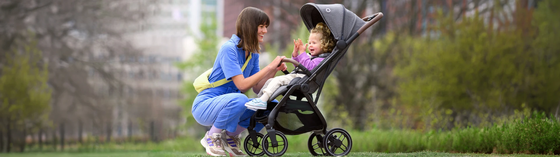 Eine Frau in Blau kniet neben einem Kinderwagen und lächelt ein Kleinkind mit lockigem Haar in einer Parkanlage an.
