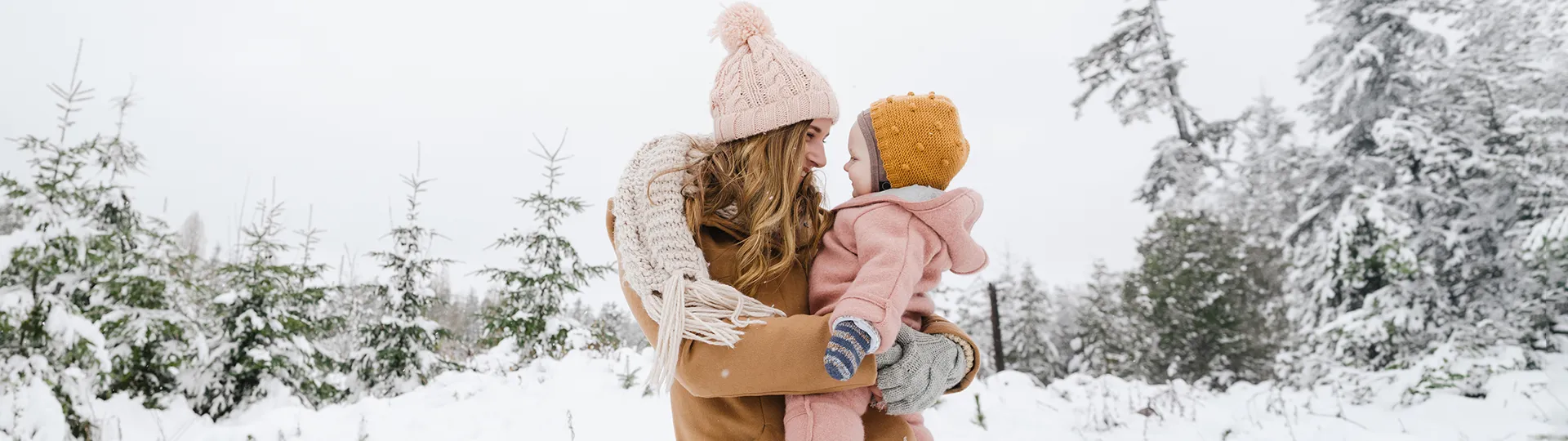 Eine Frau hält ein Kleinkind im Arm in einem verschneiten Wald; beide sind warm eingepackt mit Mützen und Mänteln und genießen gemeinsam einen freudigen Moment.