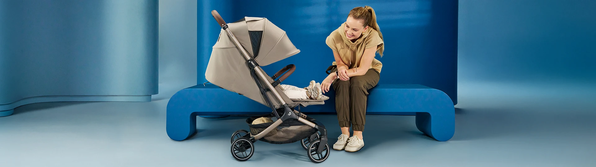 Eine Frau sitzt auf einer blauen Bank und lächelt ein Baby in einem beigen Kinderwagen vor einem modernen blauen Hintergrund an.