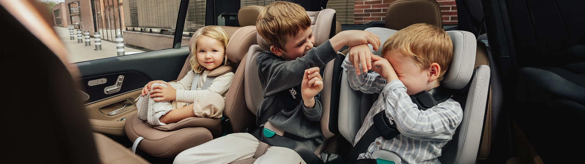 Drei Kinder sitzen in Kindersitzen. Zwei Jungen spielen miteinander, während ein Mädchen zufrieden in die Kamera lächelt.
