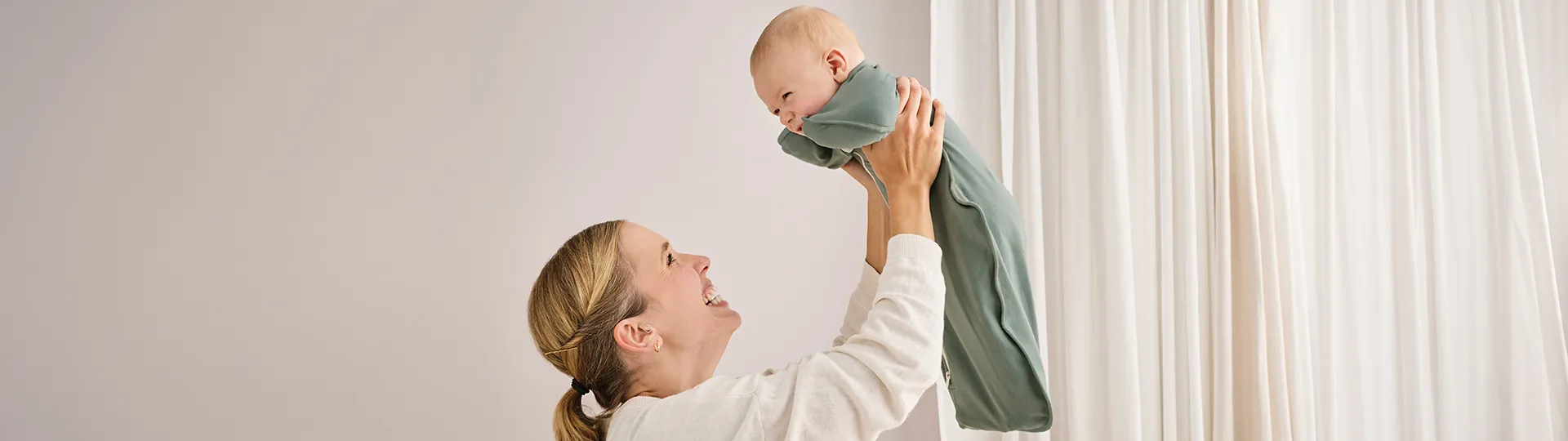 Baby wird hochgehalten und trägt einen grünen Schlafsack von Love to Dream. Der Schlafsack bietet eine bequeme Passform und unterstützt einen komfortablen Schlaf für Babys.
