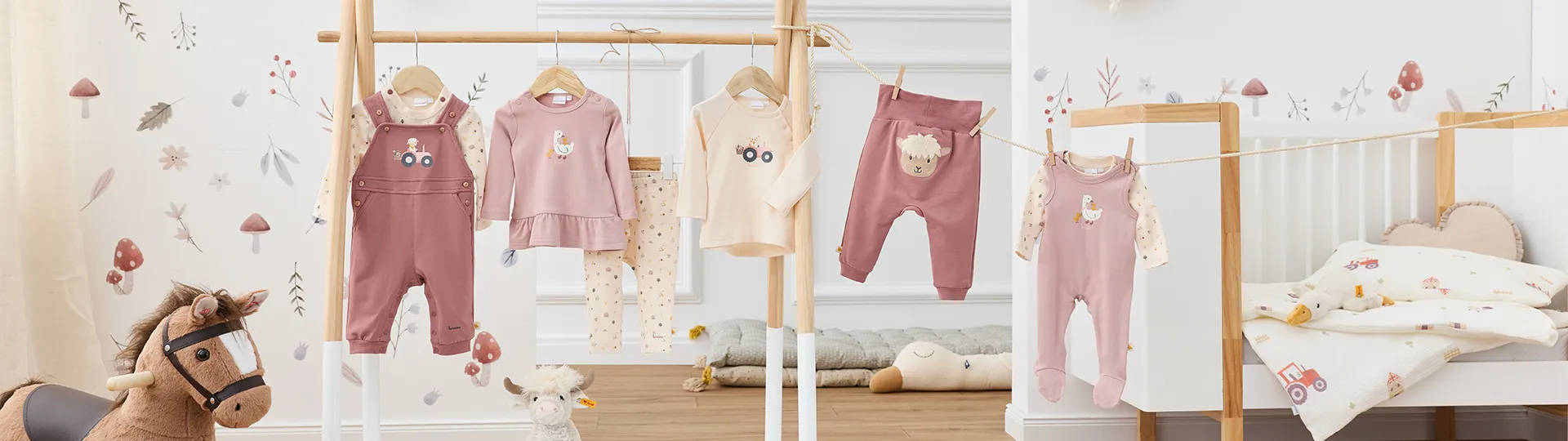 In einem Kinderzimmer mit Plüschtieren und verspielter Tapete hängt eine Sammlung von Babykleidung in zarten Pastellfarben an einem Holzständer und einer Wäscheleine.