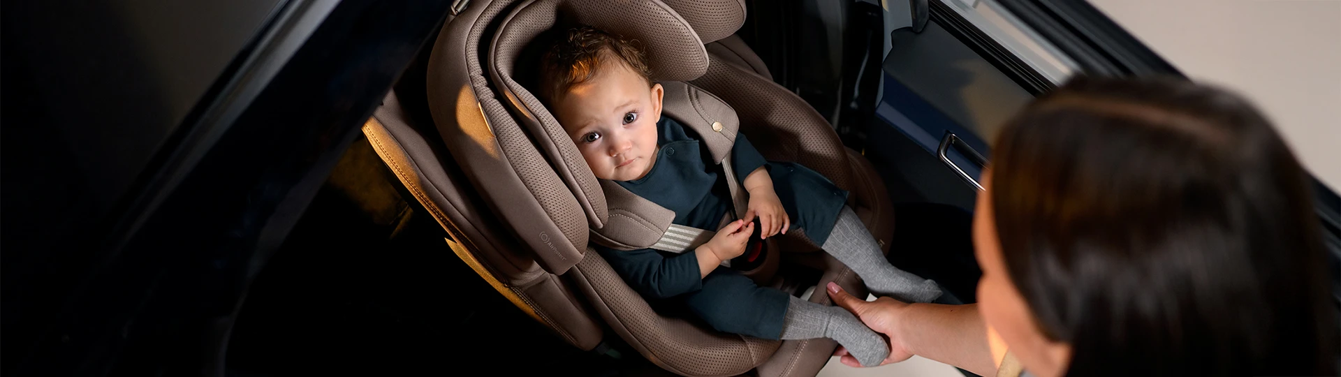 Ein Baby in einem Kindersitz blickt zu einem Erwachsenen auf, der seinen Fuß hält. Der Kindersitz ist auf dem Rücksitz eines Fahrzeugs installiert.