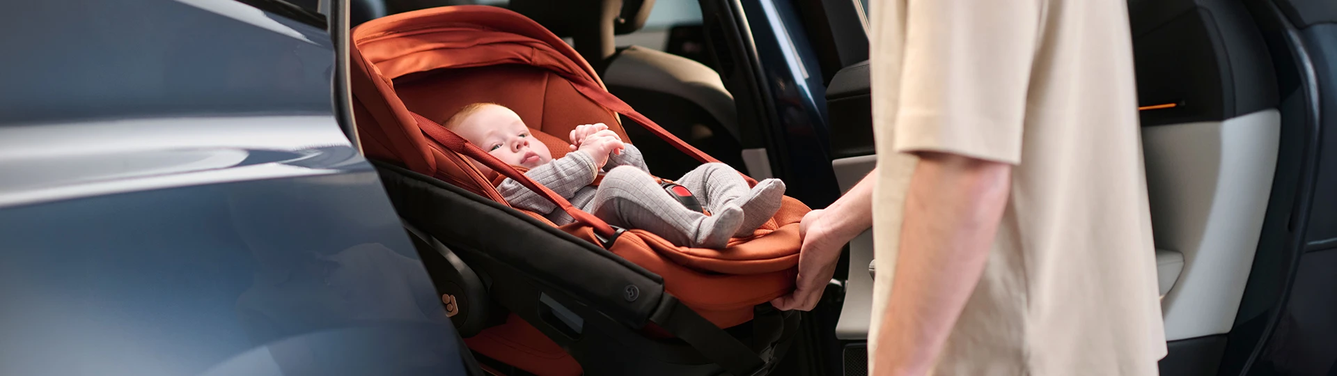 Eine Person setzt ein Baby in einem roten Kindersitz in ein Auto. Das Baby trägt graue Kleidung und wirkt entspannt und geborgen.