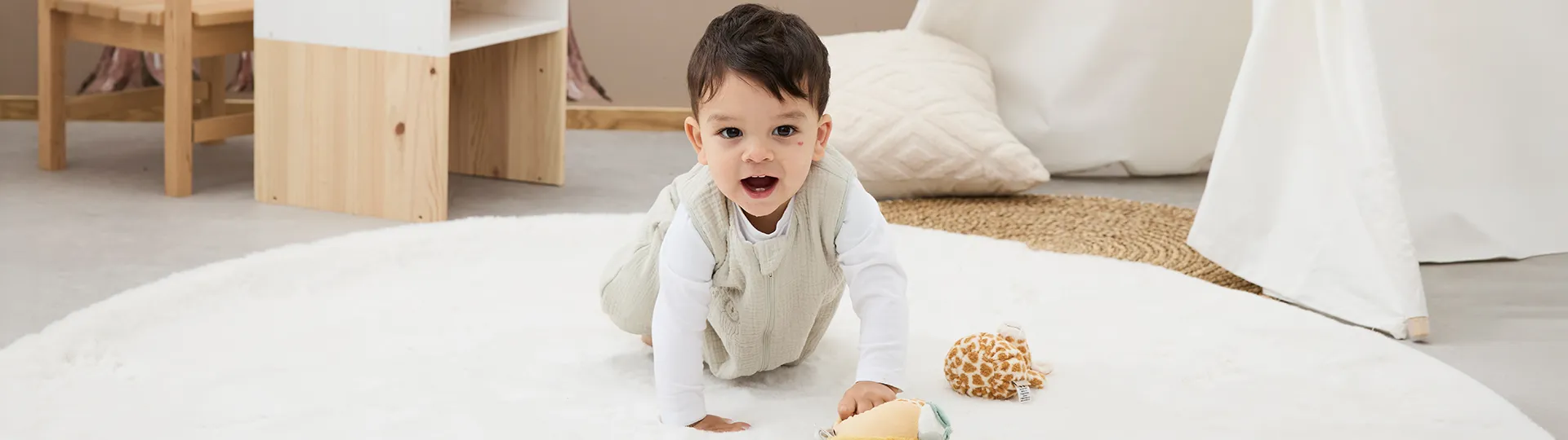 Krabbelndes Baby in hellem Musselin-Schlafsack über weißem Langarmshirt auf einer weichen Spielmatte. Vor dem Baby liegen ein gelbes Kuschelspielzeug und weitere Spielzeugelemente in einer hellen Wohnumgebung.