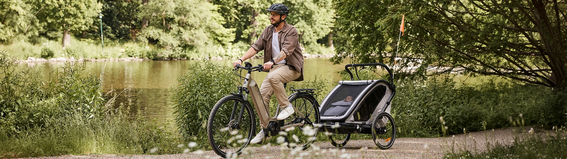 Eine Person fährt an einem sonnigen Tag mit dem Fahrrad und einem Kind in einem Anhänger auf einem malerischen Weg an einem See entlang, umgeben von üppigem Grün.