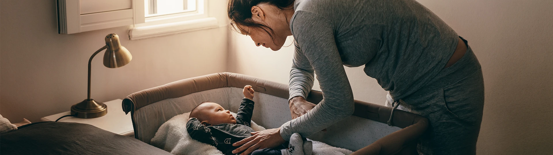 Eine Frau in grauem Pyjama beugt sich über ein Babybettchen, um sich um ein Baby zu kümmern; auf einem Beistelltisch steht eine Lampe im sanften Licht eines Fensters.