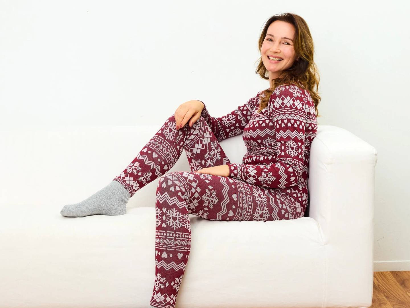 Eine Frau in einem festlichen roten Schlafanzug mit weißem Schneeflockenmuster sitzt auf einem weißen Sofa, lächelt; darunter befindet sich ein Holzboden.