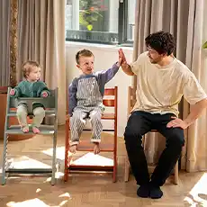 Ein Mann und zwei Kinder sitzen auf Stühlen, das ältere Kind gibt dem Mann ein High-Five, in einem hellen Raum mit Holzboden.