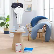 Ein gemütliches Zimmer mit einem Kissen, Kleidung auf Kleiderbügeln, einem kleinen Tisch, Handtüchern und Pflanzen als Dekoration neben einem großen Fenster.