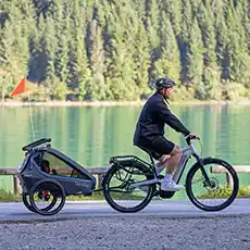 Eine Person fährt mit einem Fahrrad mit Kinderanhänger in der Nähe eines ruhigen Sees und Waldes, trägt einen Helm und Shorts.