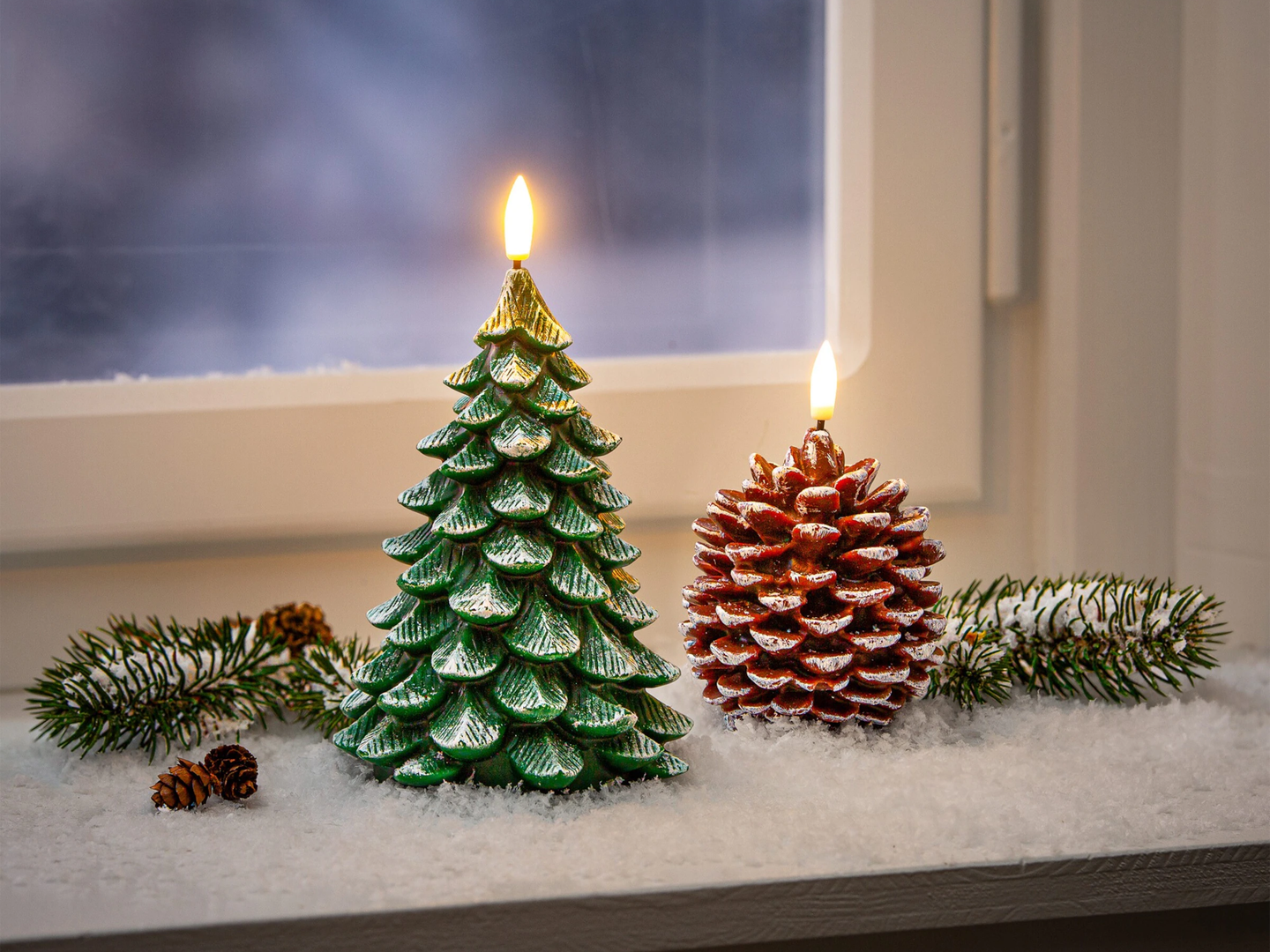 Zwei Kerzen in Form eines Weihnachtsbaums und eines Tannenzapfens auf einem verschneiten Fensterbrett, daneben Tannenzweige.