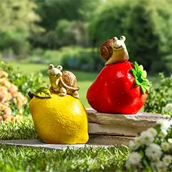 Zwei Schneckenfiguren sitzen auf großen Obstskulpturen – die eine auf einer Zitrone, die andere auf einer Erdbeere – inmitten einer üppigen Gartenlandschaft.