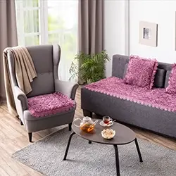 Gemütliches Wohnzimmer mit einem grauen Sessel, lila Kissen auf dem Sofa, einem kleinen Couchtisch mit Tee und Snacks sowie einem Teppich auf dem Holzboden.