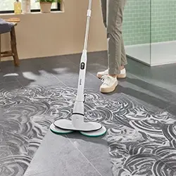 Eine Person benutzt eine weiße Bodenreinigungsmaschine auf einem dunklen Fliesenboden und hinterlässt einen sauberen Weg.