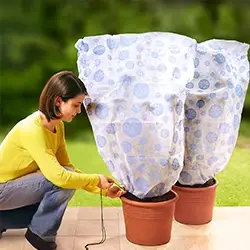 Person befestigt eine helle Pflanzenschutzhaube mit blauem Muster über zwei Topfpflanzen auf einer Terrasse vor grünem Gartenhintergrund.