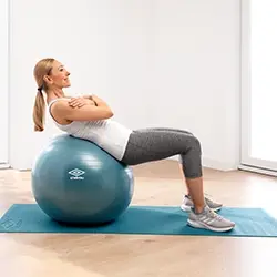 Eine Frau in Trainingskleidung macht Sit-ups auf einem blauen Gymnastikball, der auf einer Yogamatte in einem hellen Raum steht.