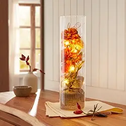 Vase mit orangefarbenen Blumen und Lichterkette auf einem Holztisch am Fenster, mit sanftem Tageslicht und herbstlicher Dekoration.