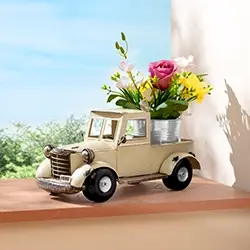 A small toy truck planter with colorful flowers sits on a ledge, with a bright blue sky and greenery in the background.