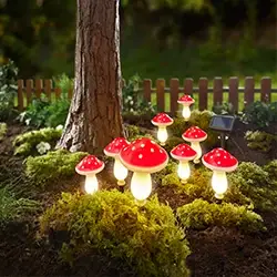 Leuchtende, pilzförmige Gartenlampen mit roten Kappen stehen zwischen Moos und Mulch in der Nähe eines Baumes, im Hintergrund ist ein kleines Solarpanel zu sehen.