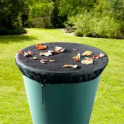 Auf einer mit Bäumen und Sträuchern umgebenen Rasenfläche steht eine Regentonne mit einer schwarzen Netzabdeckung, auf der Herbstblätter verstreut liegen.