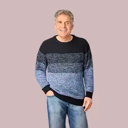 Ein lächelnder Mann in einem blau-schwarzen Pullover mit Farbverlauf und Jeans steht vor einem hellrosa Hintergrund.