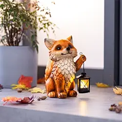 Auf einem Tisch, umgeben von Herbstblättern und Eicheln, steht eine dekorative Fuchsfigur, die eine brennende Laterne hält. Im Hintergrund steht eine Topfpflanze.