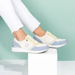 Person in weißen Hosen und stylischen Sneakers mit blauen, beigen und weißen Farbblöcken, sitzend vor einem blaugrünen Hintergrund.