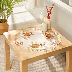 Ein Holztisch mit einer dekorativen Tischdecke im Herbstdesign mit Kürbissen und Blättern, auf dem zwei Vasen mit roten Beeren stehen, in einem hellen Raum.