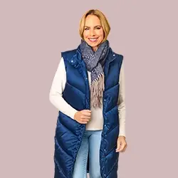 Eine Frau in einer blauen Daunenweste, einem weißen Pullover und einem Schal lächelt vor einem hellrosa Hintergrund.