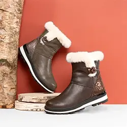 Paar knöchelhohe Stiefeletten in Dunkelbraun mit hellem Schaftrand und dekorativer Schnalle vor warmfarbigem Hintergrund mit Holzdekoration.
