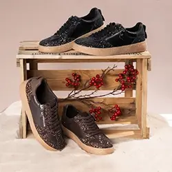 Zwei Paar Freizeitsneaker, schwarz und braun, auf einer Holzkiste mit roten Zierbeeren, vor einem zartbeigen Hintergrund.