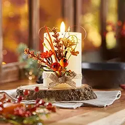 Eine brennende Kerze, verziert mit Herbstblumen und Beeren auf einem Holzsockel, steht auf einem Holztisch neben einem Fenster.