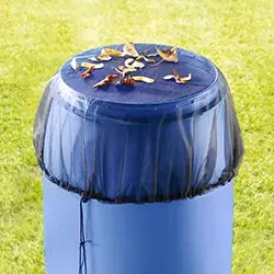 Auf einer Rasenfläche steht eine Regentonne mit schwarzer Netzabdeckung, auf der mehrere Herbstblätter liegen.