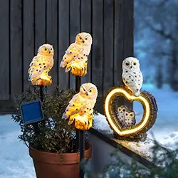 Vier beleuchtete Eulenfiguren und eine herzförmige Leuchtdekoration mit Eulenmotiv auf einer Fensterbank; daneben steht eine Eule auf einem Pflanzgefäß mit kleinem Solarpanel.