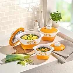 Drei orange-weiße Kochtöpfe mit Blumenmustern stehen auf einer Küchenarbeitsplatte und sind mit Gemüse gefüllt. Daneben liegen frische Kräuter und ein Lauch.
