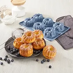 Blaue Silikonbackformen und frisch gebackene Mini-Kuchen mit Puderzucker und Blaubeeren, perfekt für kreatives Backen in der Küche.