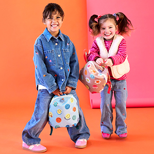 Zwei lächelnde Kinder in Jeans und bunter Kleidung halten Rucksäcke mit Smiley-Gesichtern und stehen vor einem orange-rosa Hintergrund.