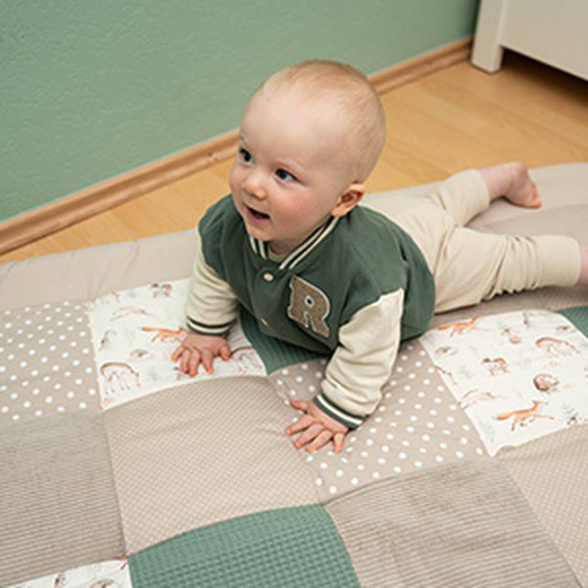 Baby liegt auf einer gepolsterten Krabbeldecke und stützt sich mit den Händen auf dem Boden ab. Die Spielmatte besteht aus verschiedenen Stoffmustern und bietet eine weiche Unterlage zum Spielen und Krabbeln.