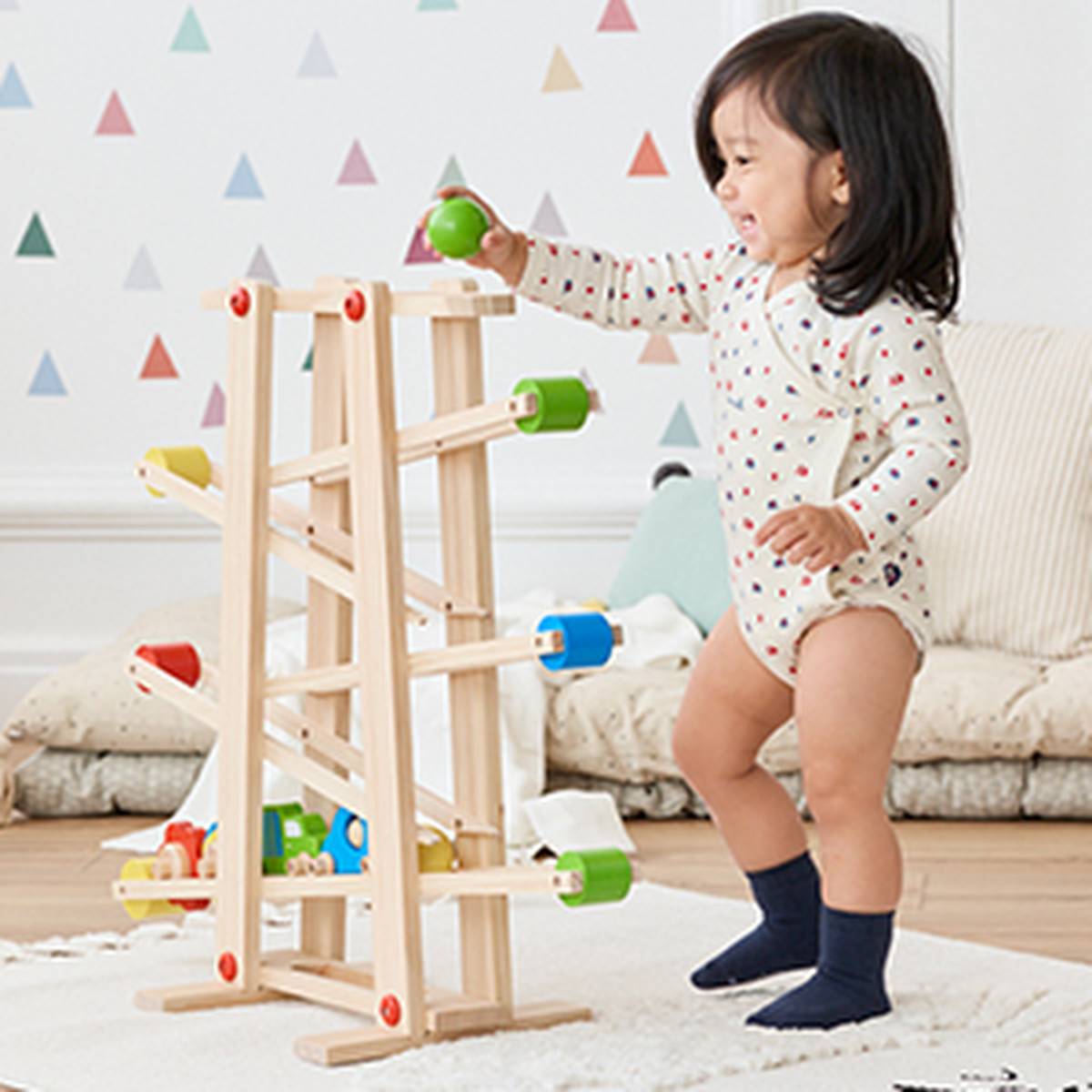 Ein Kleinkind in einem gepunkteten Strampler spielt lächelnd mit einer hölzernen Kugelbahn und hält einen grünen Ball in einem Zimmer mit dreiecksmusterigen Wänden.
