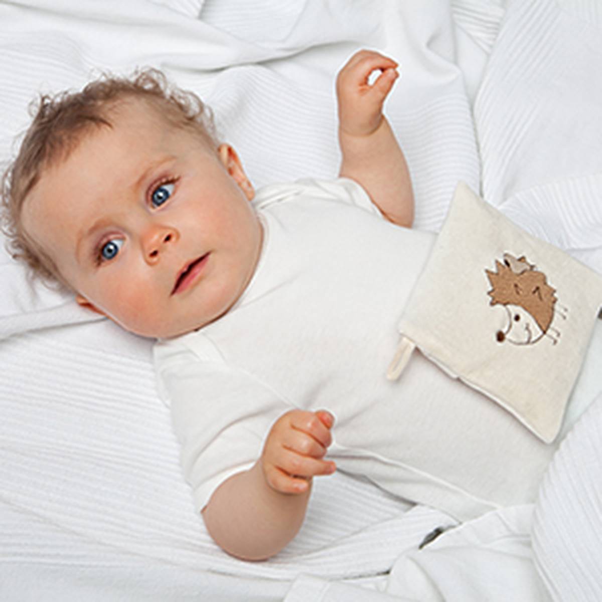 Ein Baby mit lockigem Haar liegt in einem weißen Strampler auf einer weißen Decke und hält ein kleines Kissen mit einem Igelmotiv in der Hand.