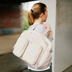 Eine Frau mit Pferdeschwanz, die eine große, beige Tasche mit mehreren Fächern trägt, steht im Freien und ist vom Betrachter abgewandt.