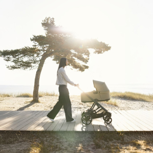 Eine Person schiebt einen Kinderwagen auf einem Holzsteg am Strand entlang, im Hintergrund sind ein einzelner Baum und strahlender Sonnenschein zu sehen.