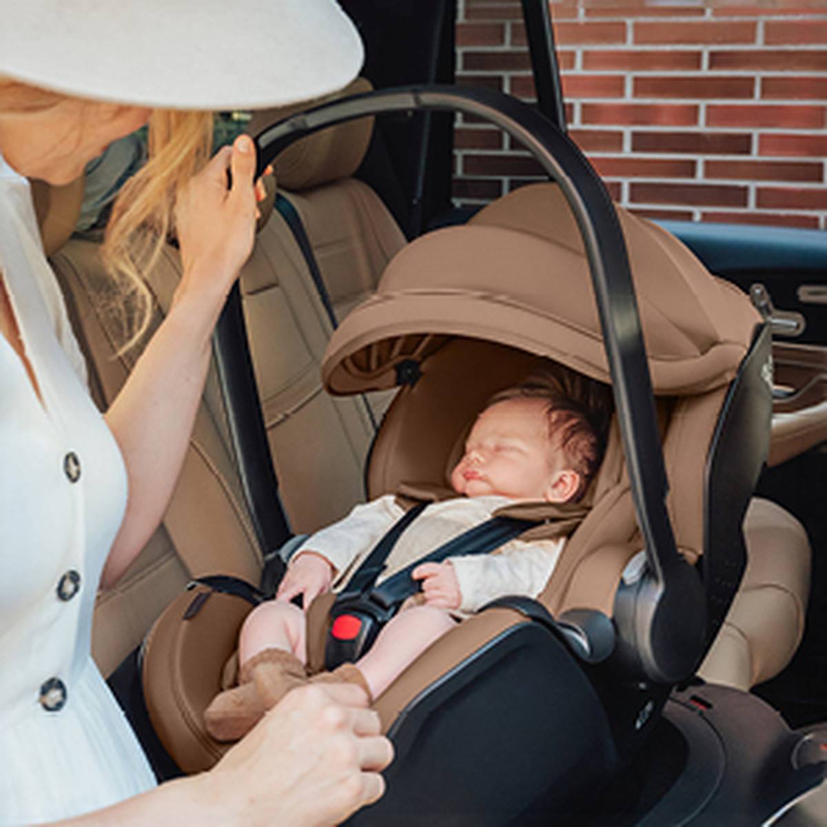 Eine Frau richtet behutsam einen Kindersitz mit einem schlafenden Baby darin zurecht, der auf dem Rücksitz eines Autos steht. Sie trägt ein weißes Kleid und einen Hut.