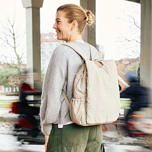 Eine lächelnde Frau in einem grauen Pullover mit beigem Rucksack steht im Freien, im Hintergrund sind verschwommen Radfahrer und Bäume zu sehen.