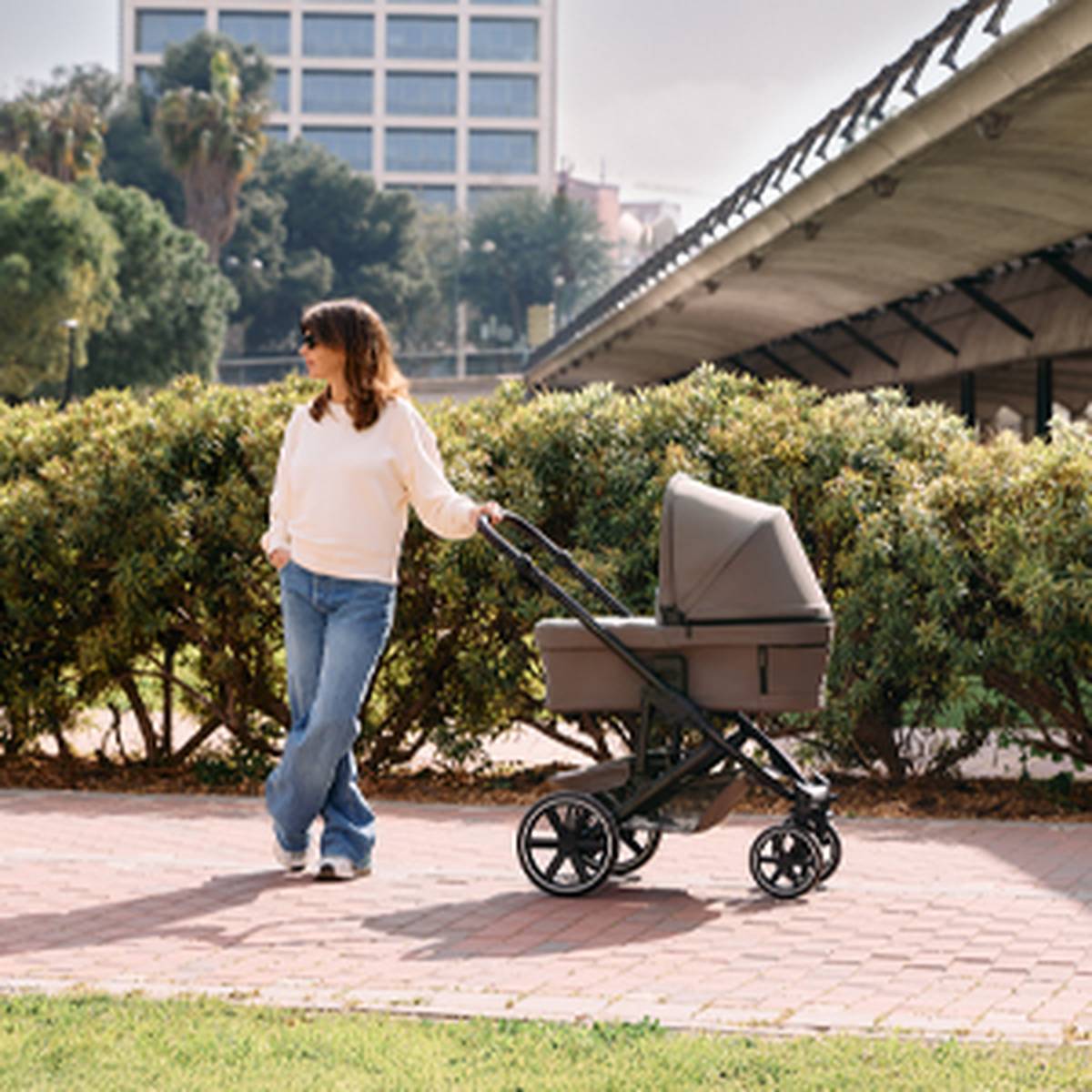 Eine Frau in einem weißen Pullover und Jeans geht mit einem Kinderwagen durch einen Park, umgeben von Grün und städtischen Gebäuden im Hintergrund.