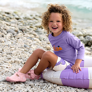 Ein lächelndes Kind mit lockigem Haar sitzt an einem felsigen Strand auf einem gestreiften Schlauchboot und trägt ein lila Shirt sowie rosa Schuhe.